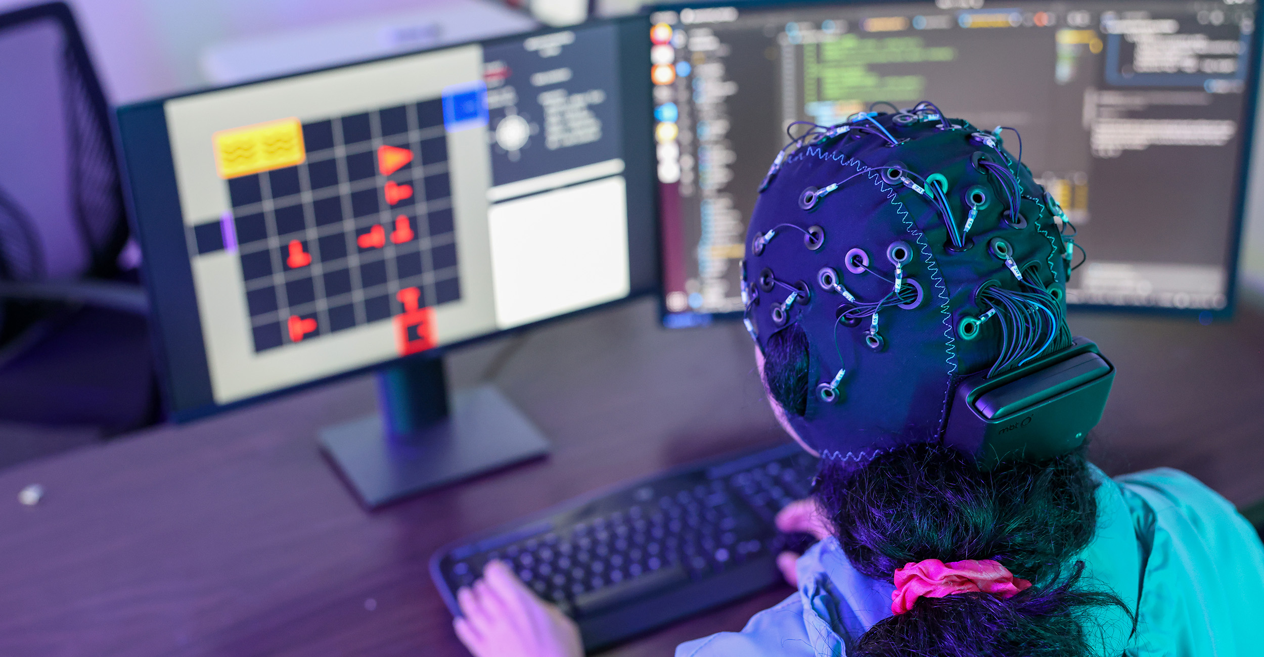 A person wearing a wired brain‑sensing cap works at a computer workstation with multiple monitors displaying data and code, illustrating neuroscience or robotics research involving real‑time brain‑computer interaction.