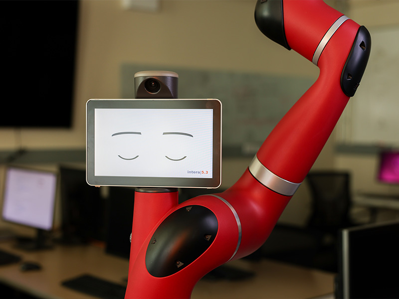 A red collaborative robot arm with a small screen displaying stylized closed eyes is positioned in a computer lab environment, illustrating a human‑friendly robotics system designed for research or interactive applications.