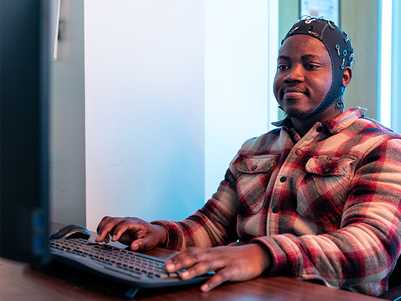 A person wearing a wired brain‑sensing cap sits at a desk typing on a keyboard while viewing code on a computer monitor, illustrating hands‑on research in brain–computer interface or robotics development.
