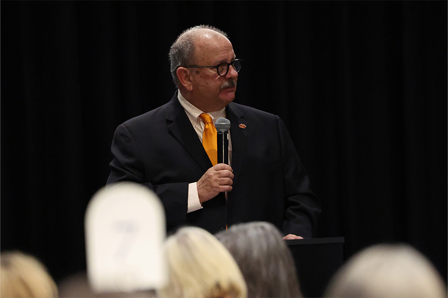 A speaker in a dark suit and orange tie stands at a podium holding a microphone and addressing an audience in a formal indoor event setting with a dark stage backdrop.