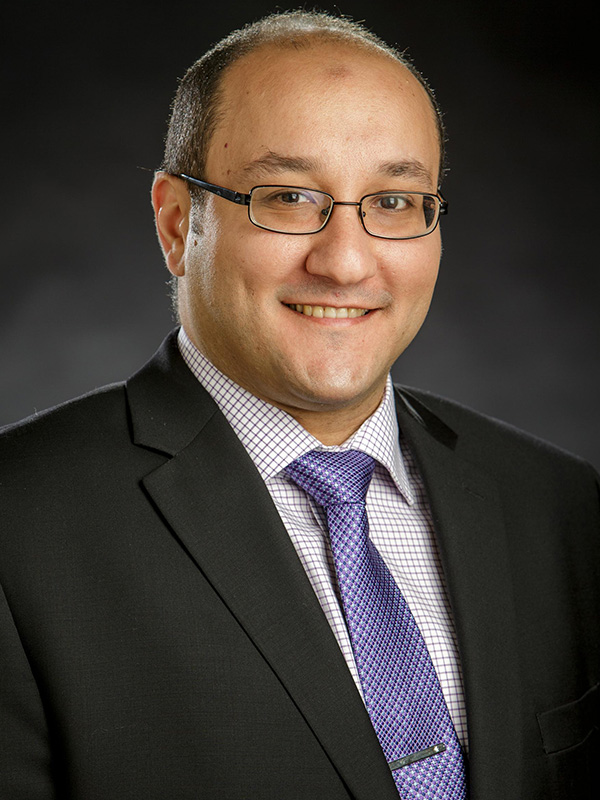 A professional portrait shows an individual wearing a dark suit, patterned dress shirt, and purple tie against a neutral studio background, conveying a formal and polished appearance.