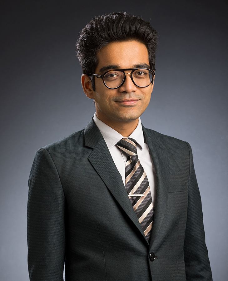 A studio portrait of a person wearing glasses, a dark suit jacket, white shirt, and striped tie against a neutral gray background.