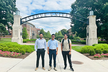 Three people standing on a walkway beneath a large “Purdue University” arch, with campus buildings, trees, and landscaped greenery on either side.