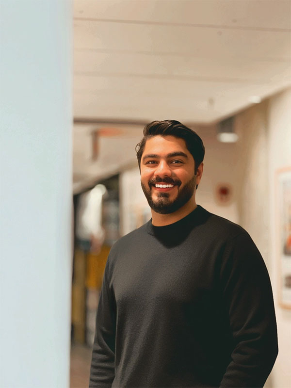 Professional portrait of an individual standing indoors in a hallway, wearing a dark sweater and facing the camera.