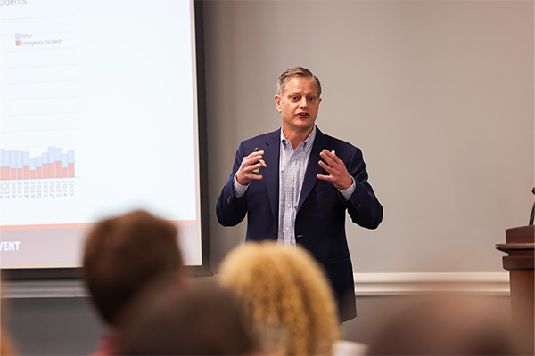 A man in a dark navy blazer and light checkered shirt stands at the front of a room, speaking and gesturing with both hands.