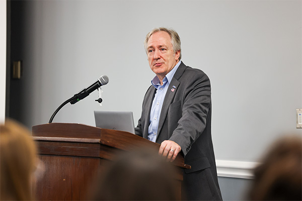 A middle-aged to older man with gray hair stands behind a dark wooden lectern in an indoor room.