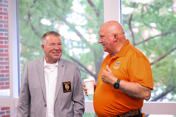 A photograph of two smiling older men standing indoors beside large windows.