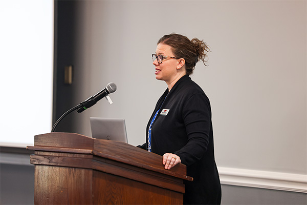 A woman stands behind a dark wooden lectern, speaking into a microphone. 