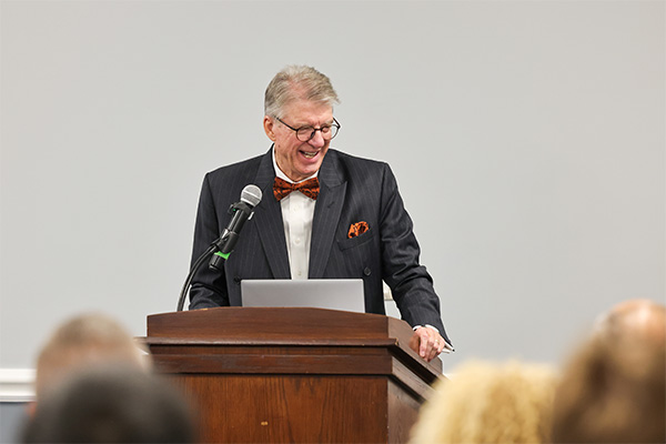 An older man stands behind a dark wooden lectern in a plain light-gray room, addressing an audience.An older man stands behind a dark wooden lectern in a plain light-gray room, addressing an audience.