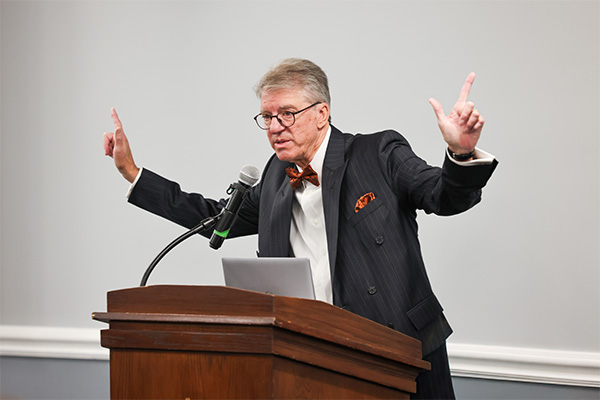 A gray-haired man wearing glasses, a dark pinstriped suit, white shirt, and orange bow tie stands behind a wooden lectern.