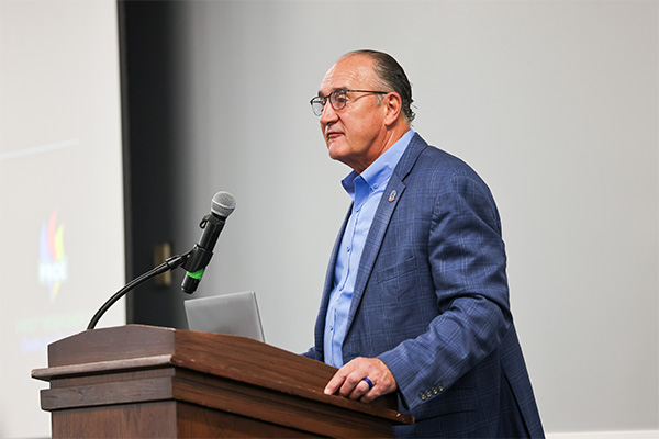 A middle-aged man wearing glasses, a blue dress shirt, and a blue checked blazer stands behind a dark wooden lectern.