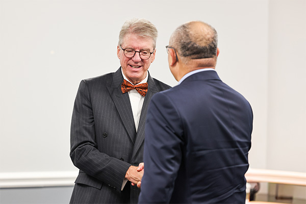 Two men stand facing each other and shaking hands in a bright, neutral-colored indoor room.