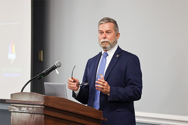 A gray-haired, bearded man in a dark blue suit, light blue tie, and white shirt stands behind a wooden lectern in an indoor presentation space.