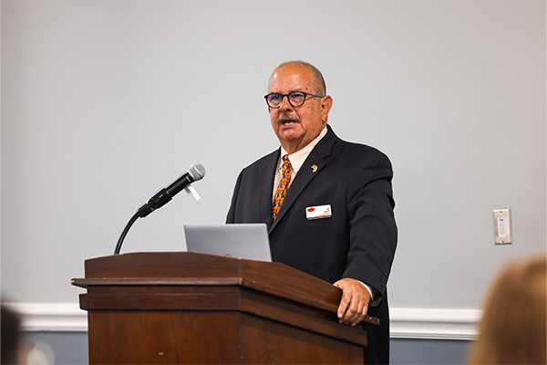 A man in a dark suit, patterned orange tie, and glasses stands behind a wooden lectern in a simple indoor room.