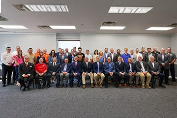 A large group of approximately 40 people pose for a formal photograph in two rows inside a carpeted conference room.