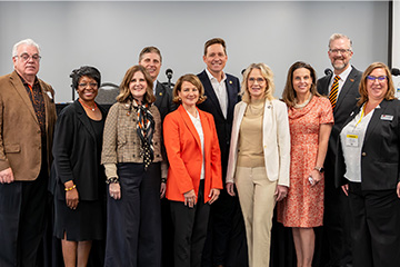 A group of professionally dressed individuals stands shoulder to shoulder indoors for a formal group photo, suggesting participation in an organized leadership, policy, or professional program.