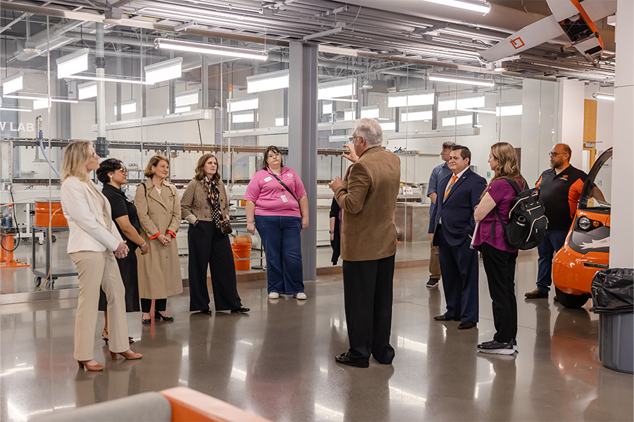 A group of professionals stands in a modern manufacturing lab while a speaker addresses them, indicating a guided tour or discussion about industrial processes and equipment.