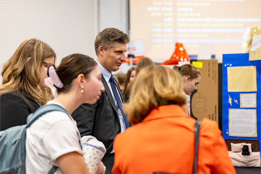 Several attendees gather closely around a student research poster display, reading and discussing the project materials during a busy academic poster session or showcase.