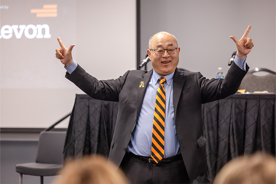 A speaker in a suit stands at the front of a conference room with both arms raised while addressing an audience, with a presentation screen and panel table visible behind him, indicating an energetic keynote or leadership talk.