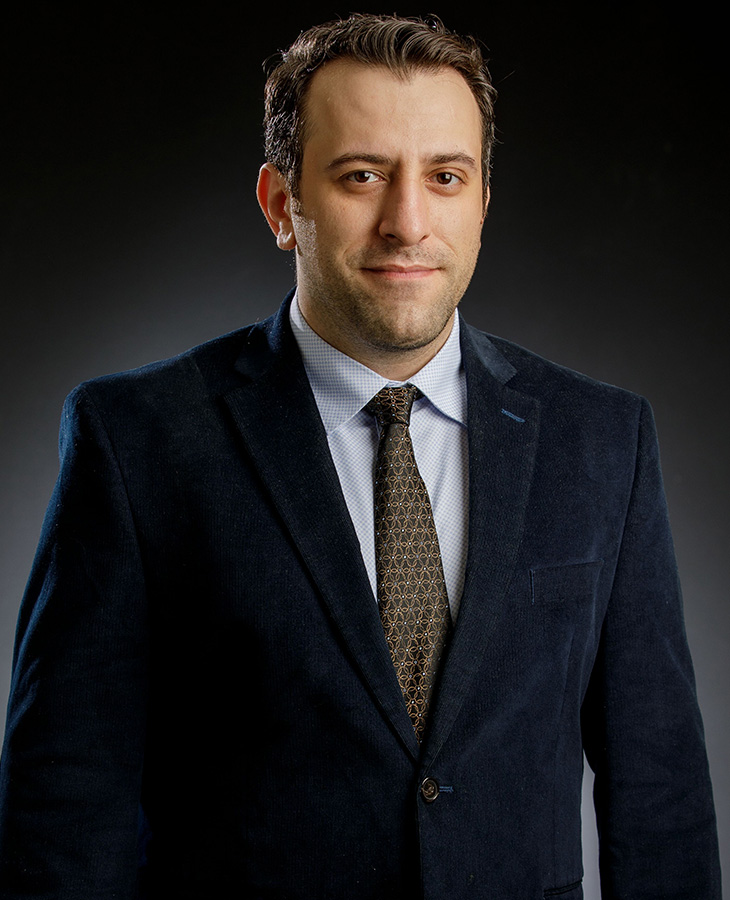 A headshot of a man in a dark suit with a multi-colored tie.