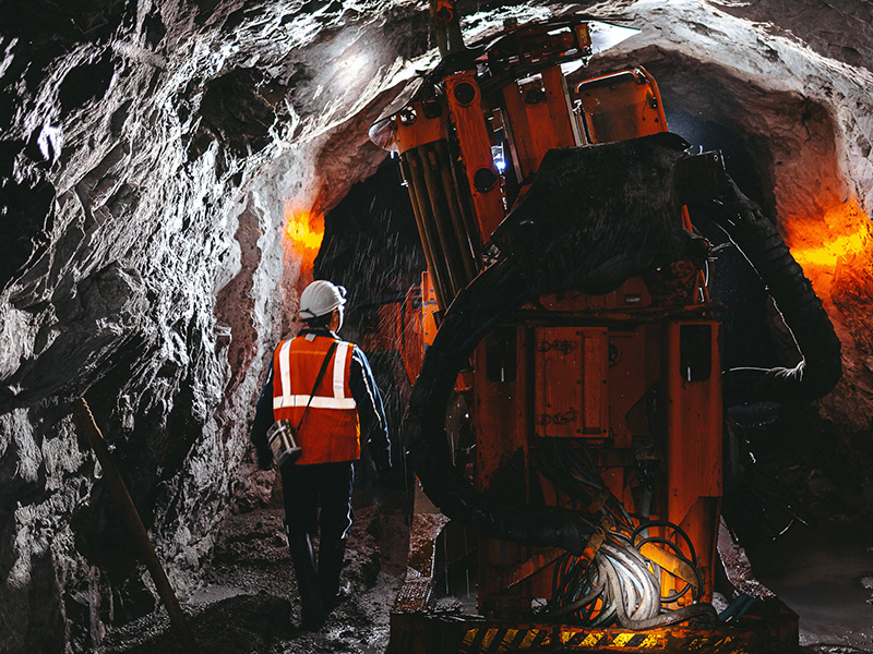 A worker wearing a hard hat and reflective safety vest walks through an underground mine tunnel beside large drilling equipment illuminated by work lights.