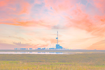 A drilling rig stands on a flat landscape near a body of water under a colorful pink and orange sunset sky.