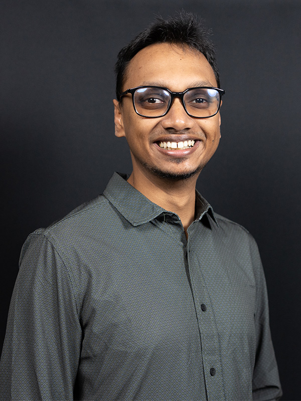 Professional headshot of a research team member wearing glasses and a dark patterned button-down shirt against a dark background.