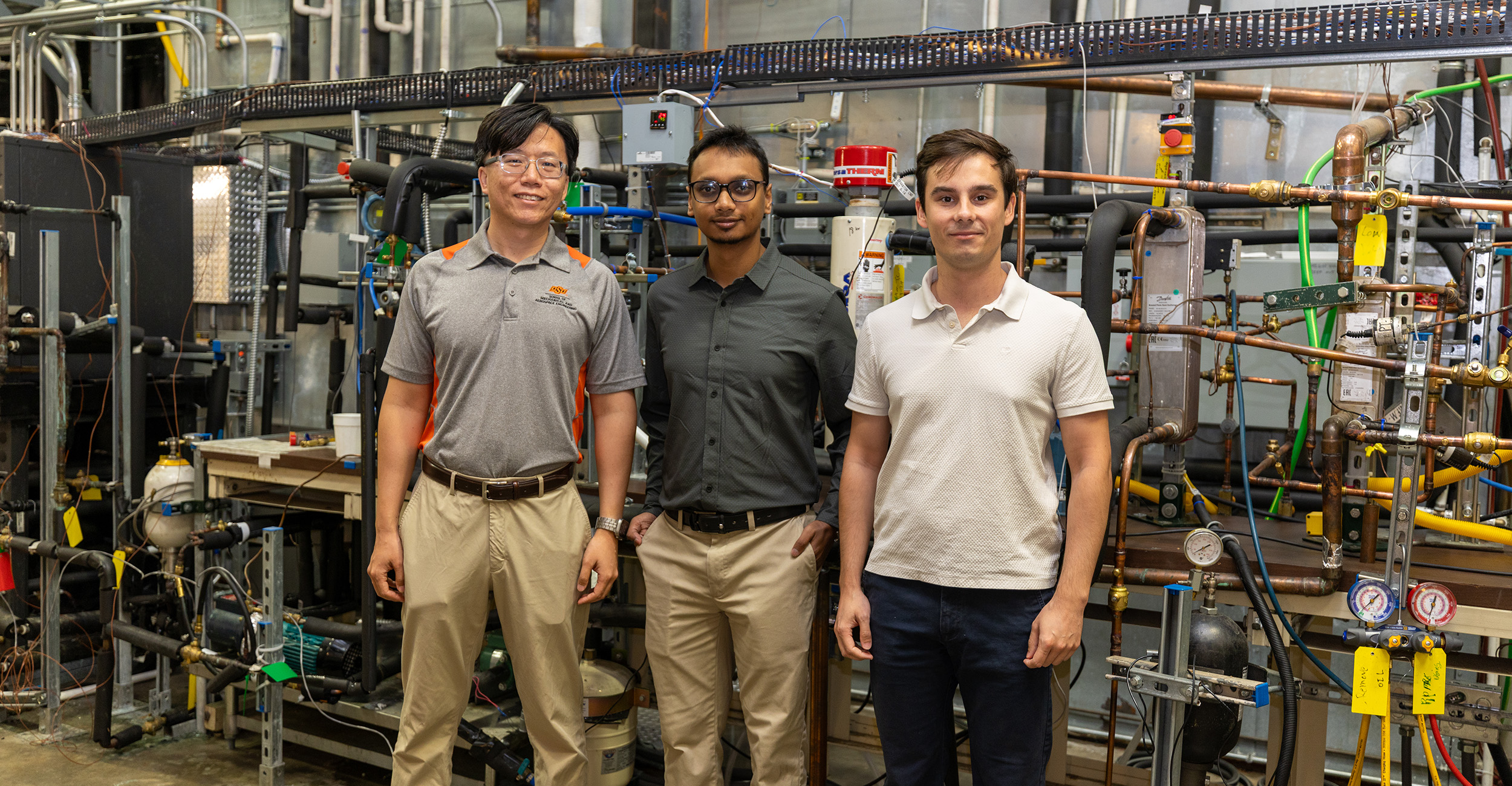 Three researchers stand in a mechanical engineering laboratory surrounded by piping, gauges, valves, and thermal systems equipment used for engineering research.
