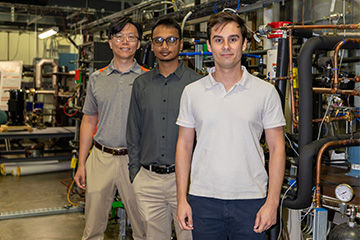 Three researchers stand in a mechanical engineering laboratory surrounded by piping, gauges, valves, and thermal systems equipment used for engineering research.