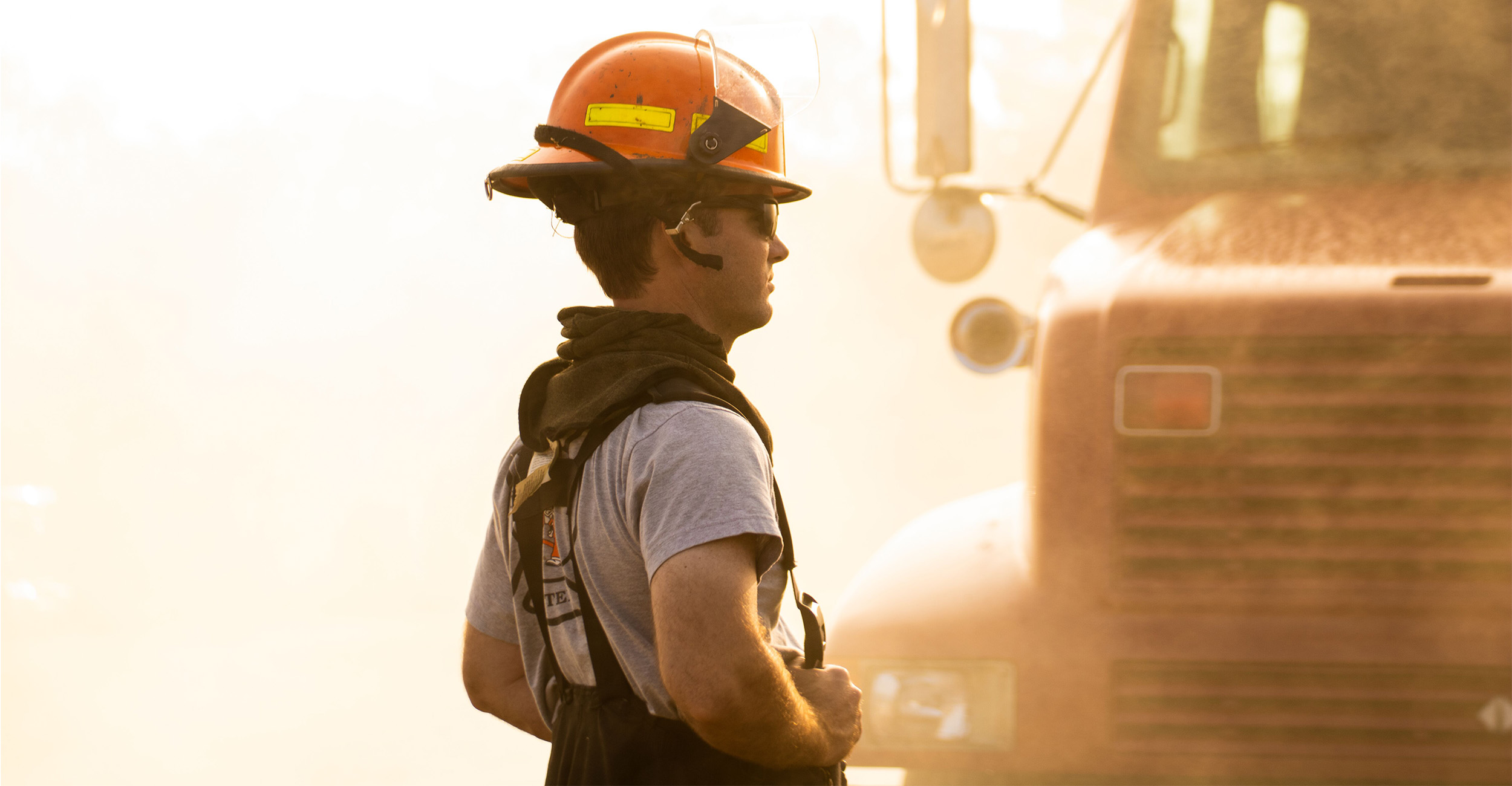 Side view of a firefighter wearing an orange helmet with attached headset and dark protective gear, standing near a fire truck partially visible in the background, with warm, smoky lighting suggesting an active fire scene.