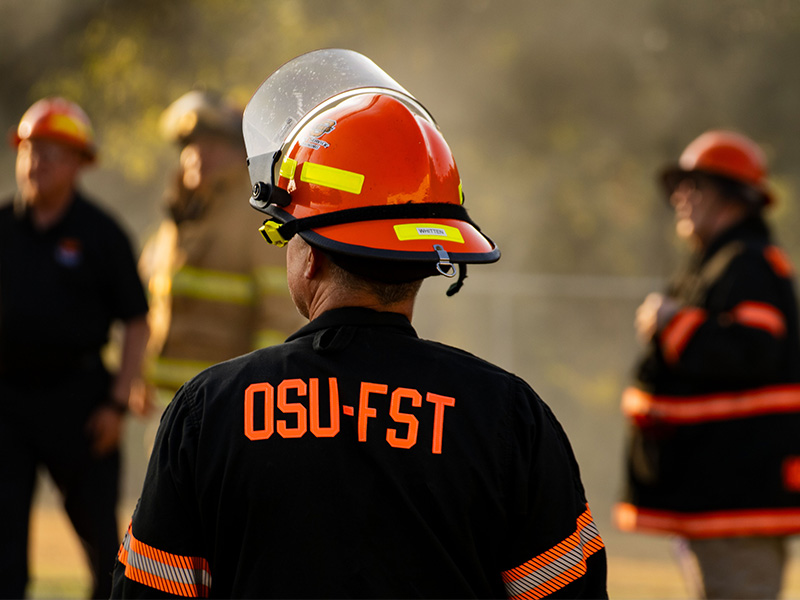 Firefighter seen from behind wearing an orange helmet with a clear visor and a black jacket labeled 'OSU-FST' in orange lettering, with several other firefighters in protective gear visible out of focus in the background.