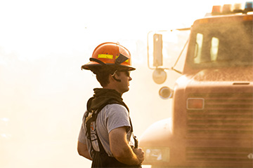 Side view of a firefighter wearing an orange helmet with attached headset and dark protective gear, standing near a fire truck partially visible in the background, with warm, smoky lighting suggesting an active fire scene.