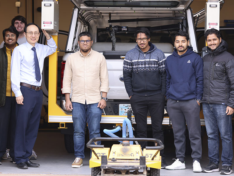 A group of researchers and students stands in front of a pickup truck outfitted with specialized equipment inside a laboratory garage. A small pavement-testing device is positioned in the foreground.