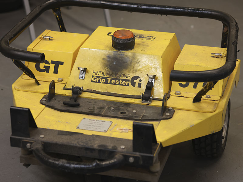 A portable yellow grip tester used to measure pavement friction and roadway surface conditions sits on a laboratory floor.