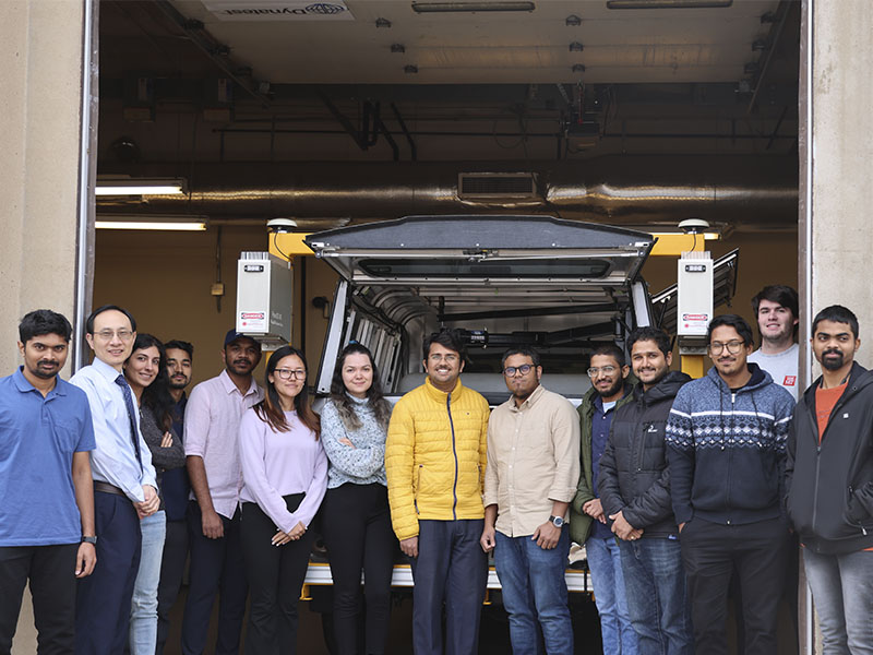A group of researchers and students stands in front of an instrumented pickup truck inside a laboratory garage used for transportation and pavement research.