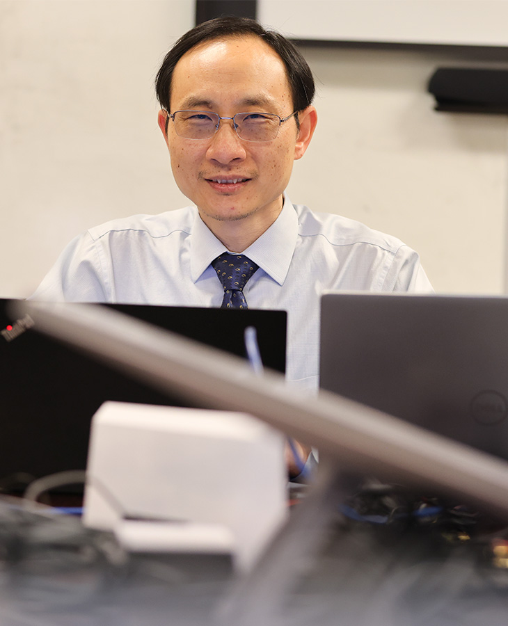 A researcher sits at a workstation surrounded by computer equipment in a laboratory setting, looking toward the camera.