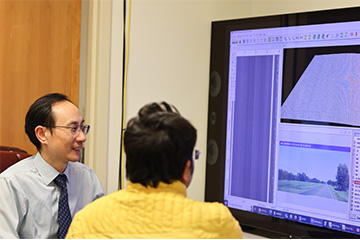 Two researchers examine pavement analysis data displayed on a large monitor. One researcher points to an image and data fields on the screen while discussing the results.