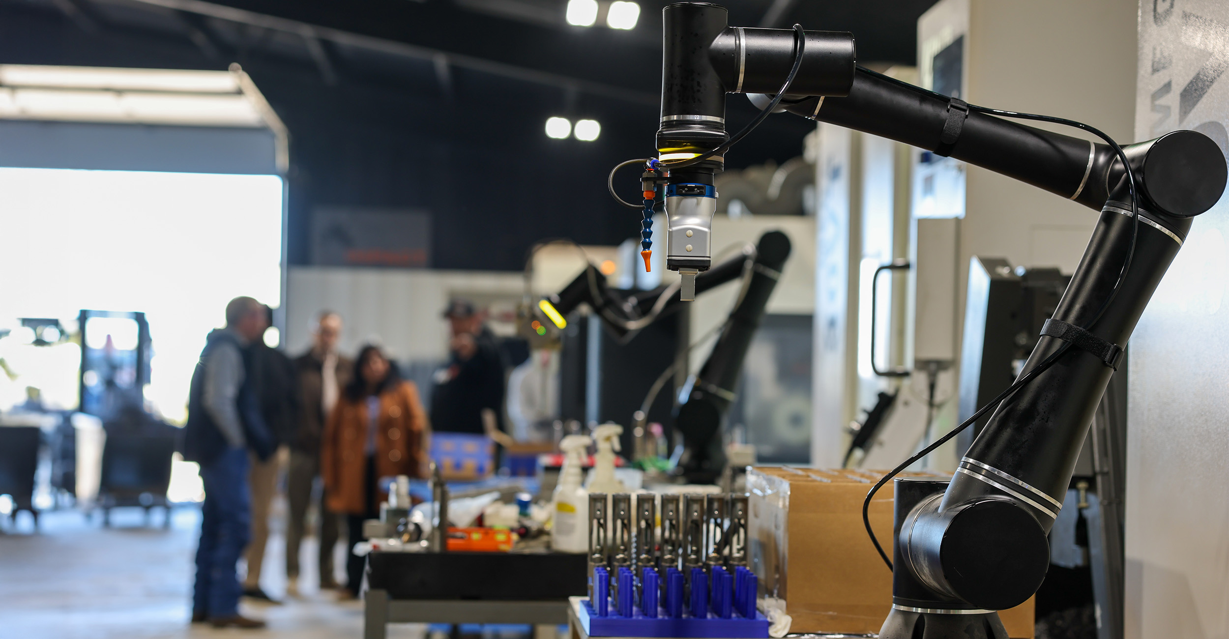 Two articulated robotic arms equipped with precision tools are displayed performing laboratory automation tasks on a workbench while attendees observe in an industrial exhibition setting.
