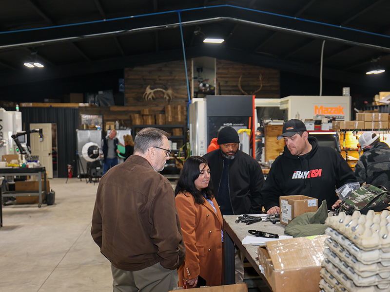 Several people stand around a workbench inside an industrial lab or workshop, observing equipment and components as part of a hands-on demonstration of manufacturing or automation processes.