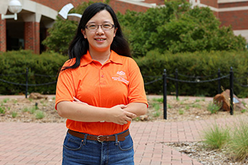 A person stands centered on a brick-paved campus walkway, facing the camera with arms crossed.