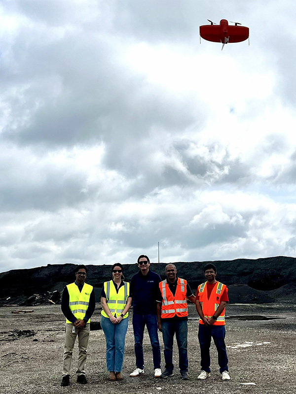 Five researchers wearing high-visibility safety vests stand at an industrial materials site while a small red drone flies overhead beneath a cloudy sky.