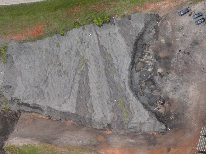 Aerial view of a large gray aggregate stockpile at an industrial site, showing surrounding work areas, vehicles, and patches of vegetation.
