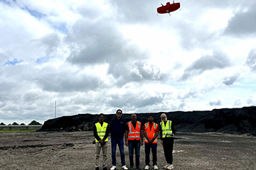 Five researchers wearing high-visibility safety vests stand at an industrial materials site while a small red drone flies overhead beneath a cloudy sky.