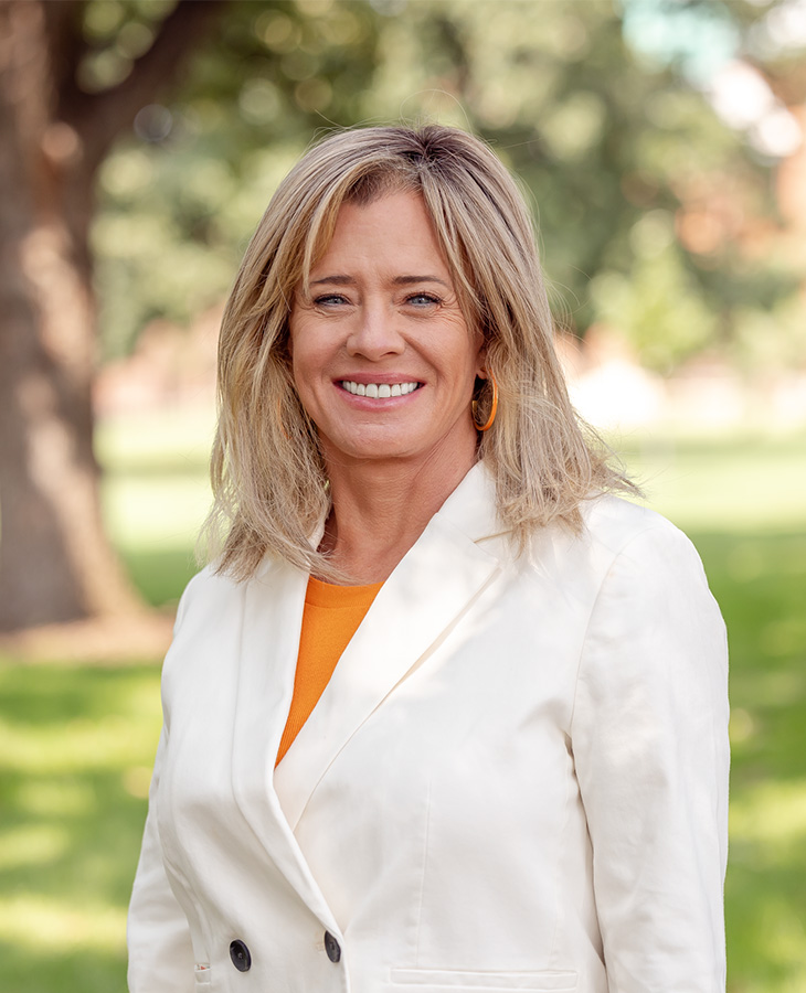 A person stands outdoors on a lawn, wearing a light-colored blazer and orange top, posing in front of trees and greenery in a professional portrait setting.