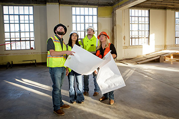 Four construction workers wearing hard hats and safety vests stand inside a building reviewing large blueprints together, illustrating collaborative planning on a job site.