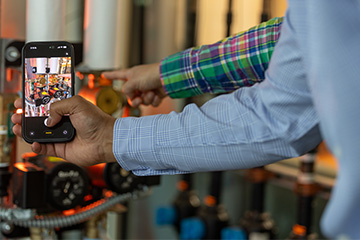 A close-up view of a person holding a smartphone in front of industrial piping equipment. The phone screen displays a photo of valves and gauges, while the person’s other hand points toward a component in the background. The setting appears to be