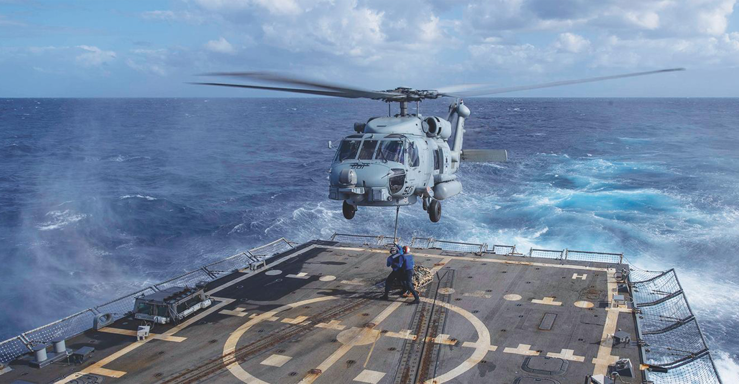 A military helicopter hovers above a ship’s marked flight deck at sea while crew members guide a cable below, illustrating a shipboard helicopter operation or resupply maneuver.