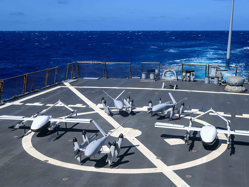 Several unmanned aerial vehicles are arranged on a marked flight deck of a ship at sea, illustrating the deployment or staging of drones for research, surveillance, or testing operations.