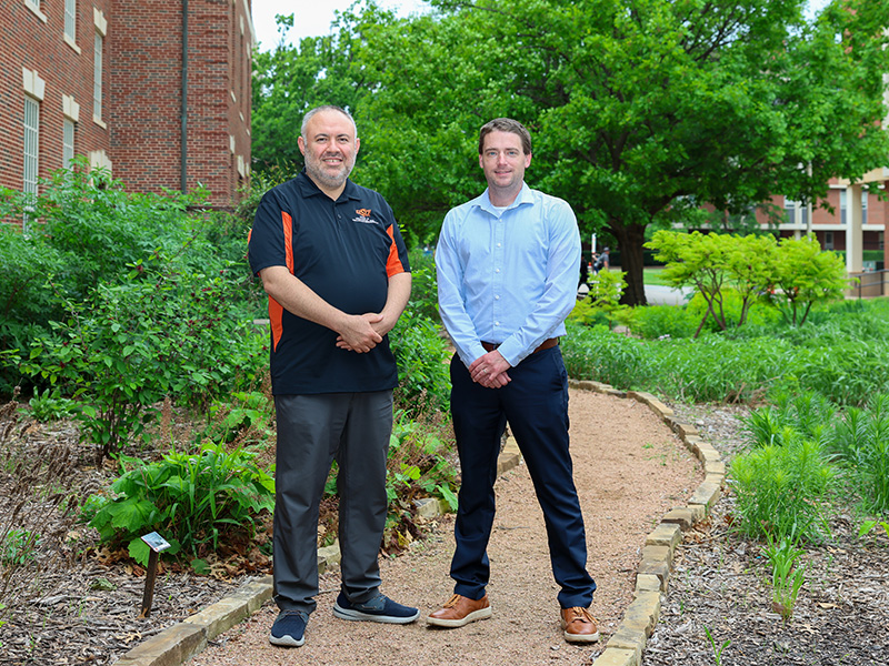 Two people stand side by side on a landscaped campus walkway with greenery and brick buildings in the background, posing for a professional outdoor photograph in an academic setting.