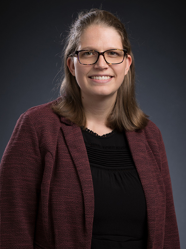 Professional studio headshot of a person wearing glasses, a dark top, and a maroon blazer, posed against a dark blue gradient background.
